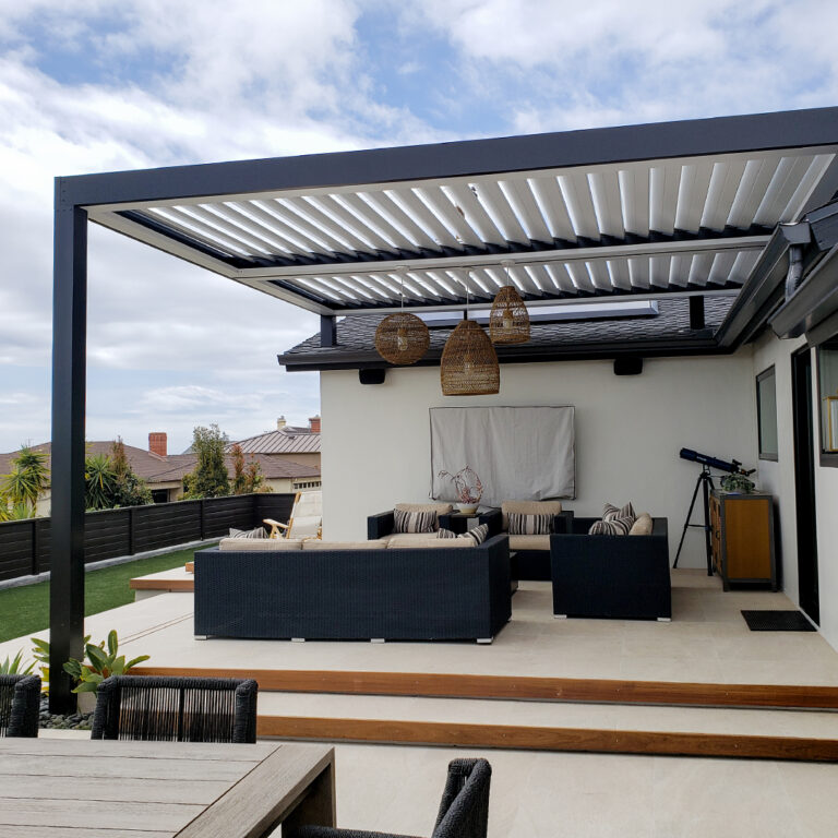 Large black aluminum louvered pergola with open louvers and hanging wicker pendant lights over a modern outdoor lounge attached to a home