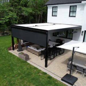 Aerial view of a black aluminum louvered pergola with a motorized privacy screen deployed on one side, covering a patio lounge and bar area beside a white farmhouse