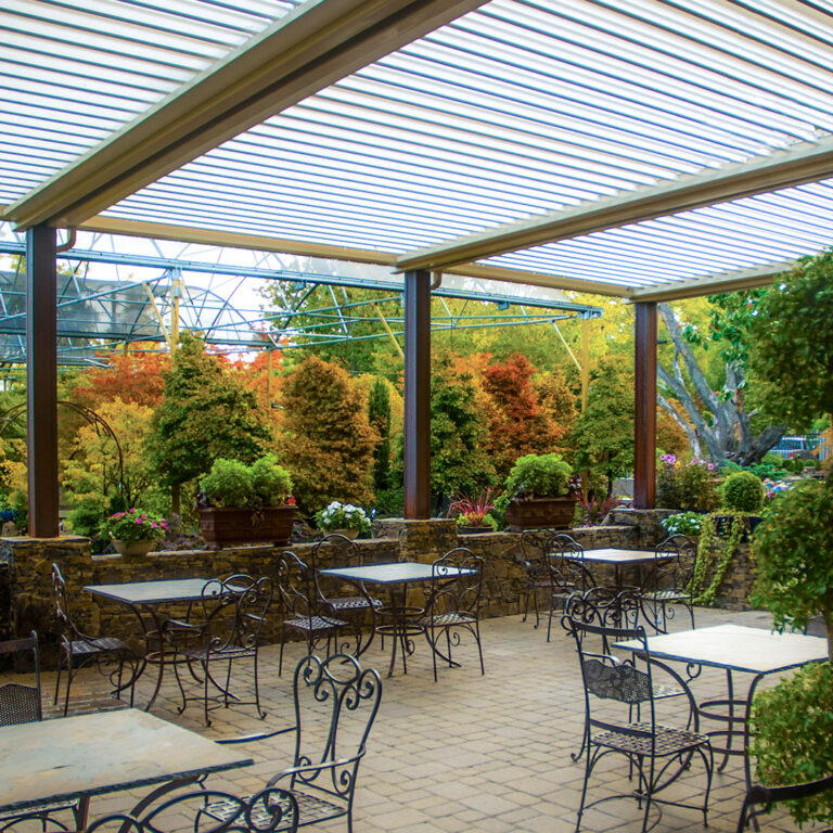 Open louvered roof over a brick patio with iron bistro tables surrounded by fall foliage and potted plants
