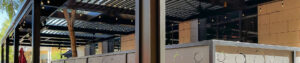 Wide-angle view of a black aluminum louvered pergola covering a restaurant patio with string lights, decorative privacy panels, and a tree-lined exterior