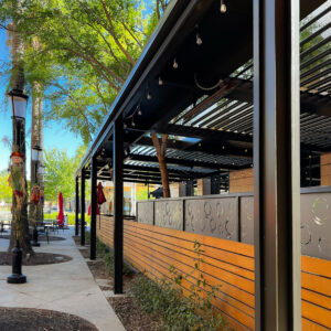Black aluminum louvered pergola covering a commercial restaurant patio with string lights along a tree-lined sidewalk