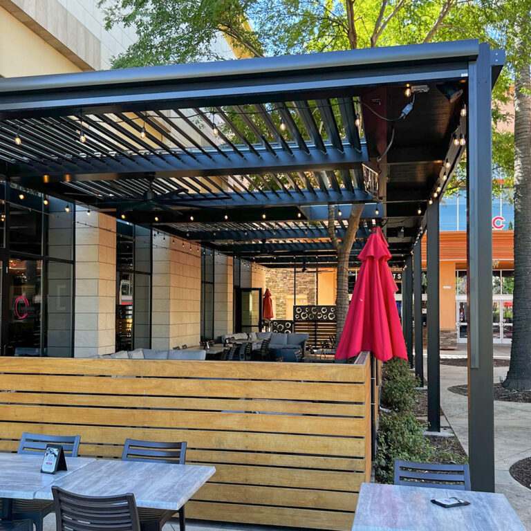 Charcoal aluminum louvered pergola spanning a restaurant patio with string lights, wood privacy wall, and open lounge seating inside
