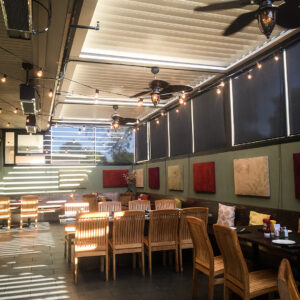 Restaurant patio fully enclosed by motorized pergola screens with ceiling fans, infrared heaters, and striped light from partially open louvers