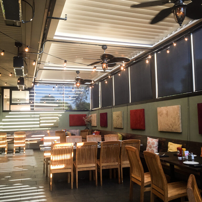 Restaurant patio fully enclosed by motorized pergola screens with ceiling fans, infrared heaters, and striped light from partially open louvers