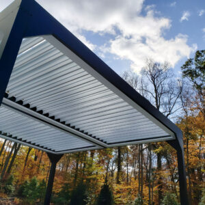 Low-angle view of the underside of a dark-framed freestanding aluminum louvered pergola with open white louvers against a blue sky, surrounded by peak autumn foliage