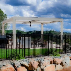 Freestanding cream aluminum louvered pergola with a ceiling fan mounted at center, installed on a patio with iron fencing and forested hills in the background