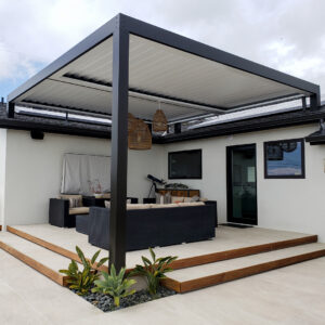 Freestanding black aluminum louvered pergola with closed louvers and wicker pendants over a raised patio with landscaped plantings at the base