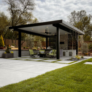 Dark freestanding aluminum louvered pergola with closed louvers, recessed lighting, and ceiling fans over an outdoor kitchen and dining area in a landscaped backyard