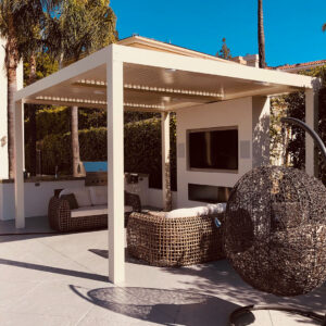 Cream aluminum freestanding louvered pergola with closed louvers and recessed speakers over a luxury outdoor lounge with a linear fireplace, outdoor TV, wicker seating, and palm trees
