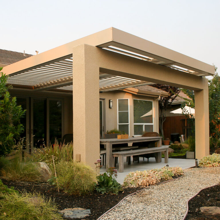 Tan freestanding aluminum louvered pergola with stucco-clad posts color-matched to a stucco home, covering a weathered wood dining set on a concrete patio with drought-tolerant landscaping at dusk