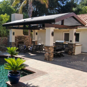 Dark aluminum gable-style louvered pergola covering an outdoor kitchen, fireplace, and seating area beside a pool with stone columns