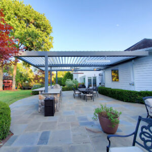 Gray aluminum louvered pergola with open adjustable louvers and mounted heaters over a stone-tile patio with outdoor dining set and built-in grill at dusk