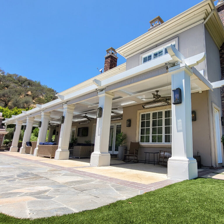 Wide white louvered pergola with decorative columns, ceiling fans, and infrared heaters spanning the full rear elevation of a large estate home