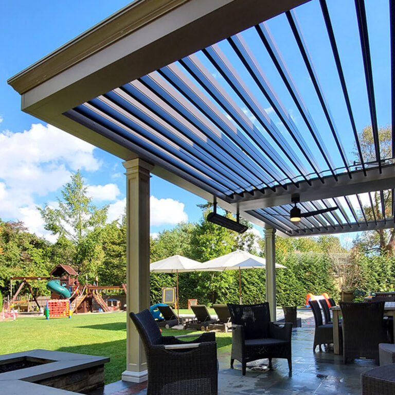 Under-canopy view of a large two-tone aluminum louvered pergola with open louvers, beam-mounted infrared heater, and ceiling fan over a flagstone patio with wicker seating, fire table, and a children's playset on the lawn beyond