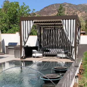 Freestanding dark-framed aluminum louvered pergola with open louvers and black-and-white striped curtains forming a poolside cabana with lounge seating and a built-in grill, backed by a mountain range