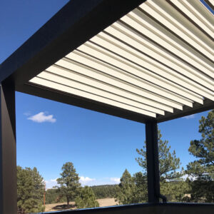 Corner view beneath a dark-framed aluminum louvered pergola with fully closed cream louvers framing a ponderosa pine forest and wide blue sky in a high-altitude Western setting
