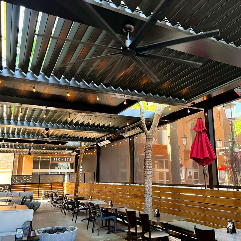 Nearly closed aluminum pergola louvers with motorized drop screens deployed, ceiling fan running, and string lights glowing over a restaurant patio