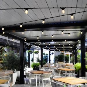 Closed aluminum louvered pergola over a garden restaurant patio with string lights, wall-mounted fans, hanging planters, and teak dining tables