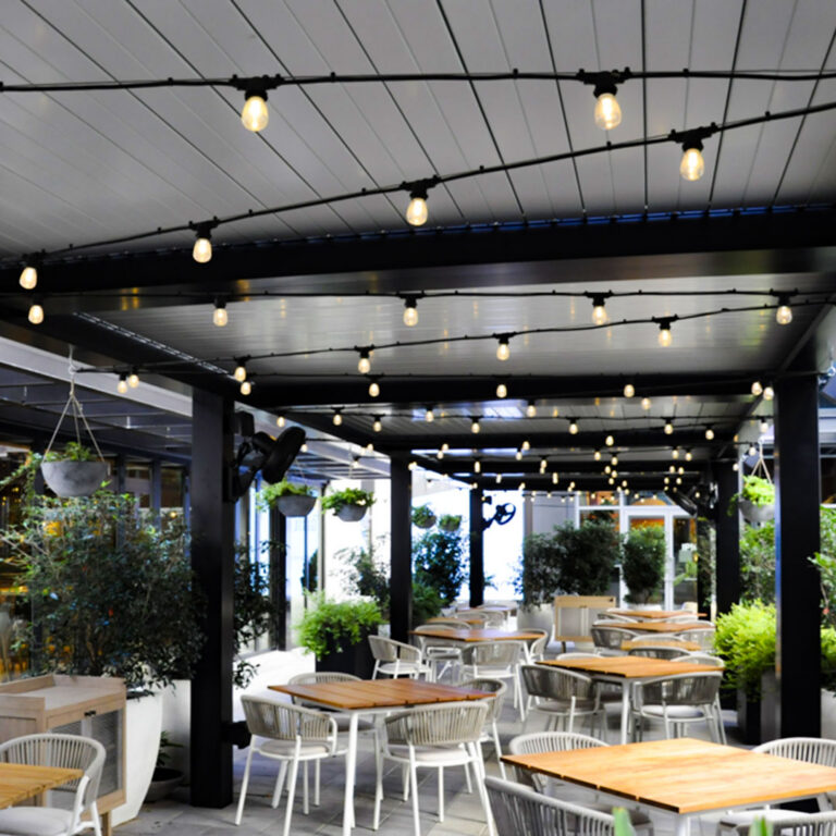 Closed aluminum louvered pergola over a garden restaurant patio with string lights, wall-mounted fans, hanging planters, and teak dining tables