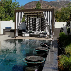Dark aluminum motorized pergola with black and white striped curtains drawn around a poolside seating area, with mountains visible behind