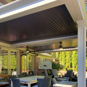 Under-canopy view of a two-tone aluminum louvered pergola with dark closed louvers, white trim frame, dual ceiling fans with lights, and an infrared heater attached to a classic brick home with French doors