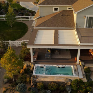 Aerial drone view of a white louvered pergola mounted on a second-story deck beside a swim spa, with rural landscaping and a white fence beyond