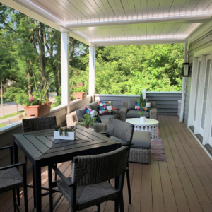 White aluminum louvered pergola with closed louvers and LED perimeter lighting over a wood deck with a bar-height dining table and wicker lounge seating surrounded by mature trees