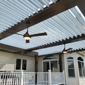 Under-canopy view of a dark-framed aluminum louvered pergola with open white louvers and two actively lit bronze ceiling fans with light kits attached to a vinyl-sided home over a white-railed deck