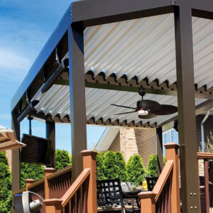 Low-angle view of a dark-framed aluminum louvered pergola with open cream louvers, individual blue-tinted point LED lights along the beam, a ceiling fan with light kit, and a wall-mounted outdoor TV on a wood deck