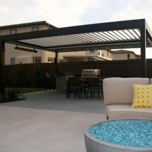 Dark aluminum freestanding louvered pergola with open louvers, wall-mounted infrared heater, and recessed lighting at dusk over a dining set and built-in grill, with a glass fire pit and lounge chair in the foreground