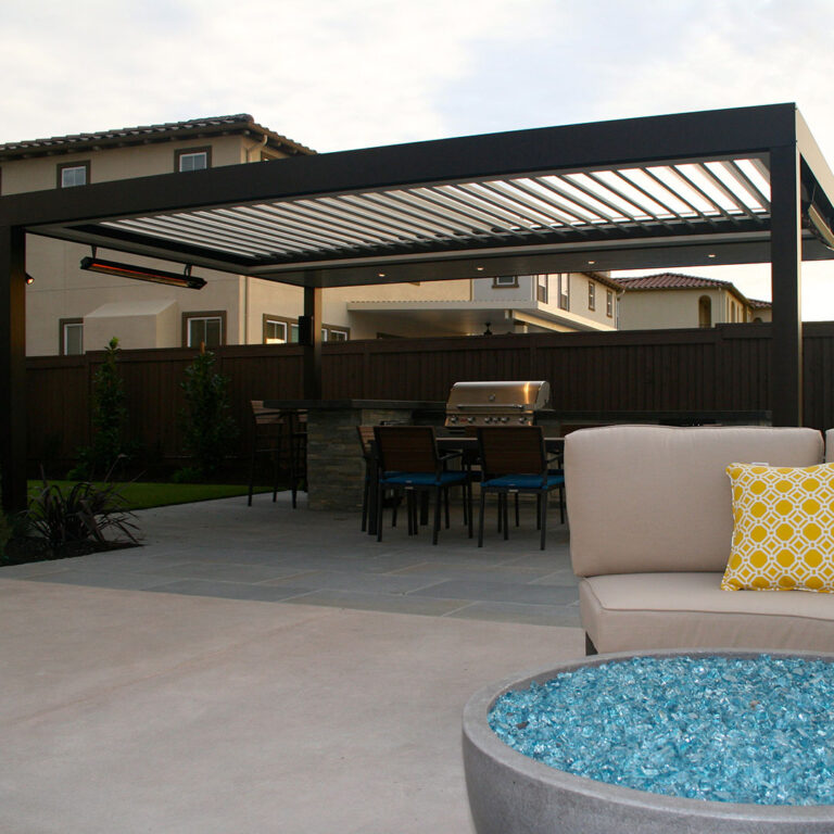 Dark aluminum freestanding louvered pergola with open louvers, wall-mounted infrared heater, and recessed lighting at dusk over a dining set and built-in grill, with a glass fire pit and lounge chair in the foreground