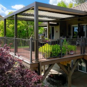 Dark aluminum louvered pergola with open louvers mounted on an elevated second-story deck with iron railing, ceiling fan, and mature trees beyond