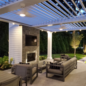 White aluminum louvered pergola illuminated at dusk with recessed lighting and string lights, overlooking a lounge area with a lit stone fireplace and wall-mounted TV