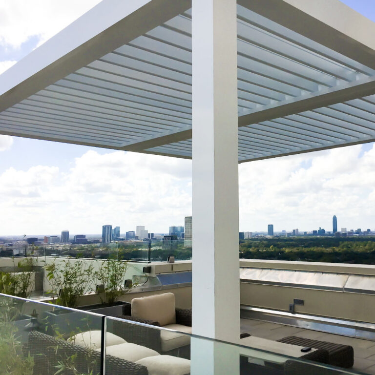 White aluminum louvered pergola with open louvers on a high-rise rooftop terrace with wicker lounge seating, glass railings, and a wide city skyline panorama