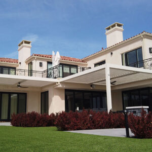 White aluminum louvered pergola with ceiling fans installed across the rear patio of a Mediterranean-style two-story home