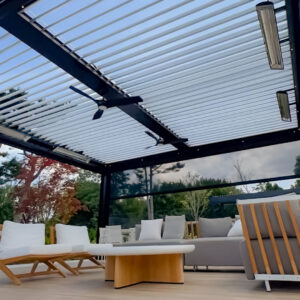 Under-canopy view of a dark aluminum louvered pergola with open louvers, motorized drop screens fully deployed, three infrared heaters, dual ceiling fans, and point LED lighting over a teak and linen deck lounge with fall foliage beyond