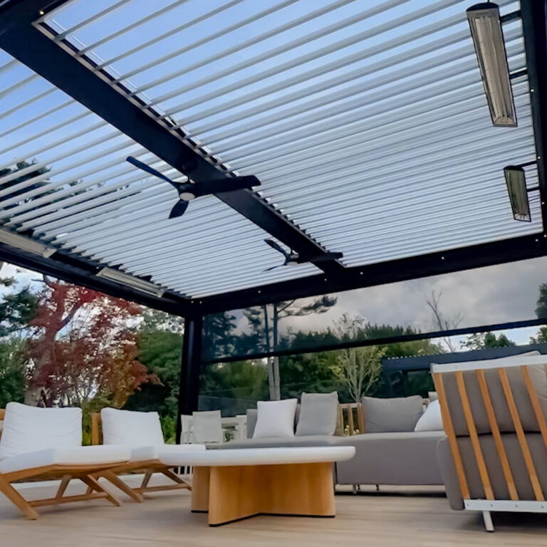 Under-canopy view of a dark aluminum louvered pergola with open louvers, motorized drop screens fully deployed, three infrared heaters, dual ceiling fans, and point LED lighting over a teak and linen deck lounge with fall foliage beyond