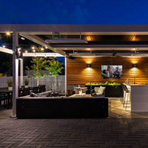 White aluminum louvered pergola at night with infrared heaters, ceiling fan, and string lights over a fire pit lounge, outdoor TV on a wood-clad wall, bar seating, and dining area on a herringbone paver patio