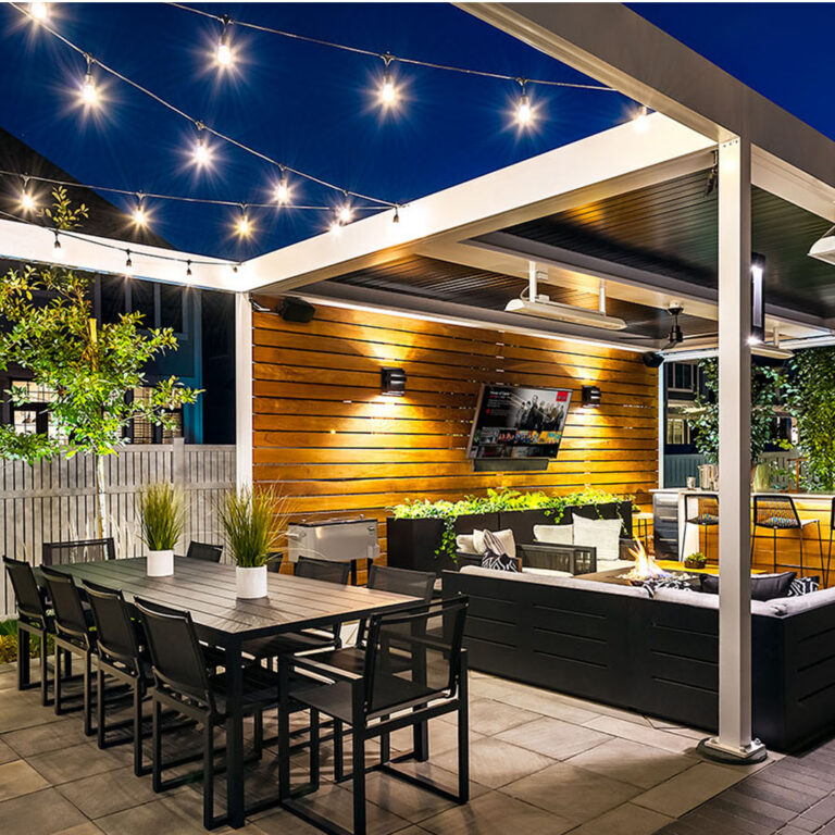 White aluminum louvered pergola with open louvers and string lights over a nighttime patio with a long dining table, fire pit lounge, outdoor TV, ceiling fan, and wood-clad feature wall