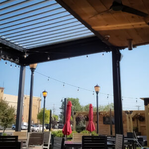 Upward view from inside a fully open louvered pergola framing a clear blue sky with street lamps and closed red umbrellas visible beyond