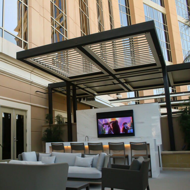Aluminum louvered pergola over a commercial outdoor bar with marble surround, wall-mounted TV, and lounge seating below