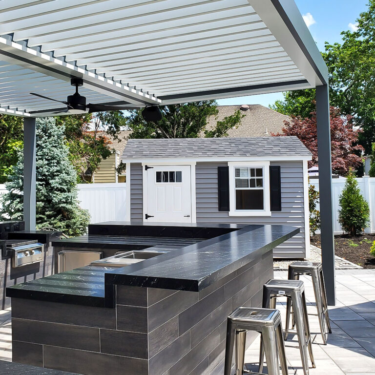 White aluminum louvered pergola with open louvers and a ceiling fan shading a dark stone outdoor kitchen bar with stainless bar stools and a built-in grill