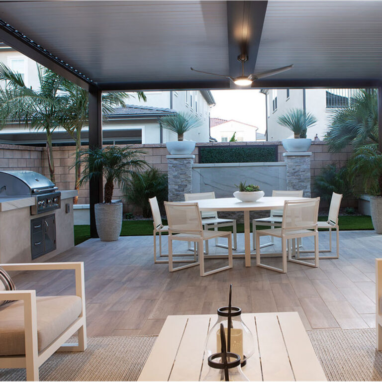 Dark aluminum louvered pergola with closed louvers, illuminated ceiling fan, and LED perimeter lighting over a modern patio with a built-in grill, round dining set, and tropical planters at dusk