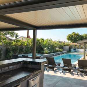 Under-view of a closed aluminum louvered roof over a dark stone outdoor kitchen counter, with wicker chaise lounges and a pool waterfall feature beyond