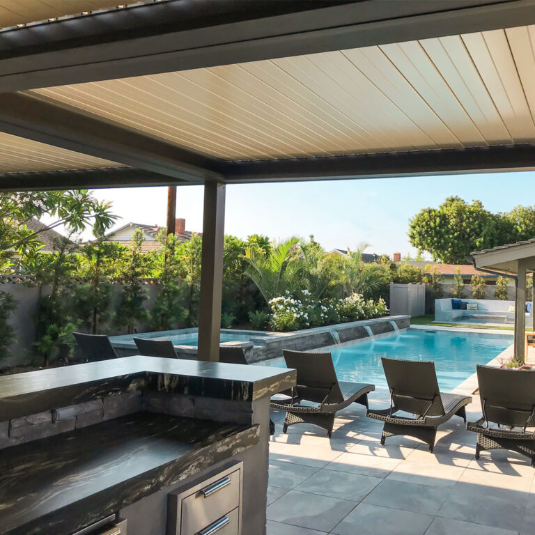 Under-view of a closed aluminum louvered roof over a dark stone outdoor kitchen counter, with wicker chaise lounges and a pool waterfall feature beyond