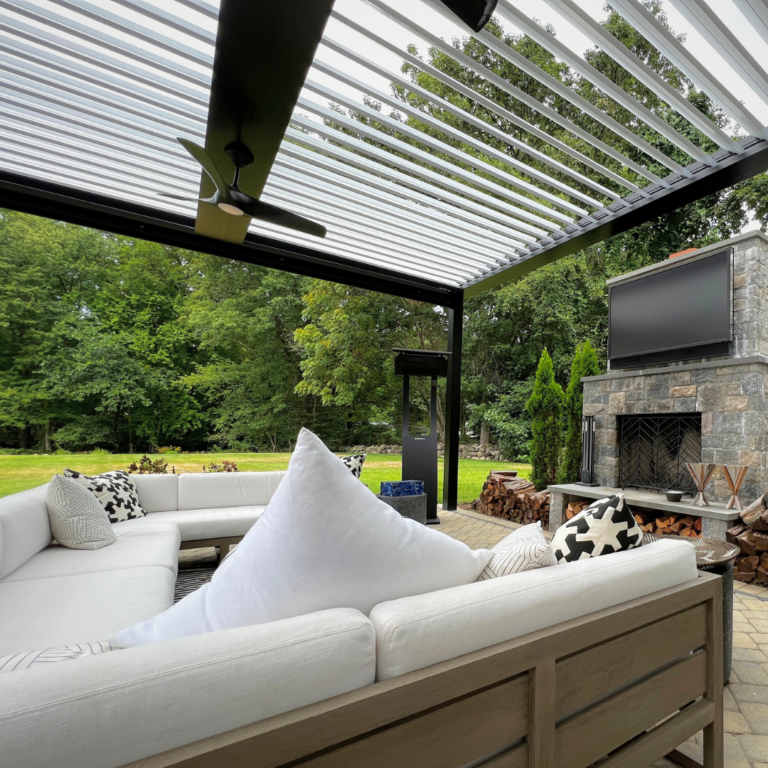 Sectional sofa and outdoor TV beneath a dark-framed aluminum louvered pergola with open louvers, a ceiling fan, and a freestanding heater beside a stone fireplace