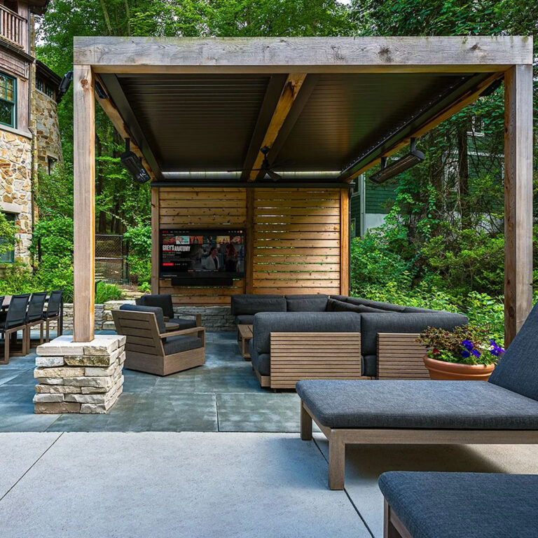 Louvered roof pergola enclosing an outdoor lounge with a mounted TV, ceiling fan, and infrared heaters among wooded surroundings