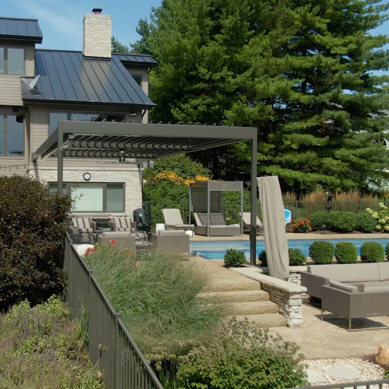 Taupe aluminum pergola with open adjustable louvers attached to a stone and shingle estate home, overlooking a landscaped pool deck with lounge chairs and garden beds