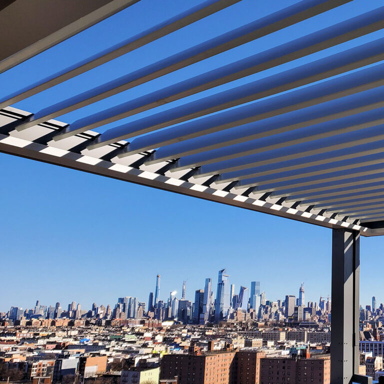Upward view through open white aluminum louvers on a high-rise rooftop terrace pergola framing the full Manhattan skyline under a clear blue sky