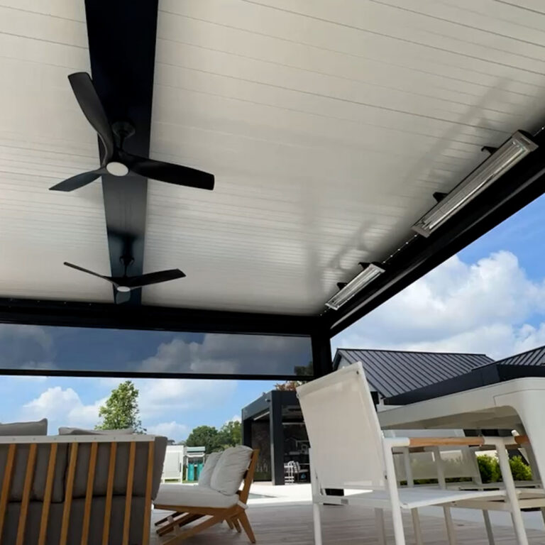 Under-canopy view of a dark aluminum louvered pergola with closed cream louvers, dual black ceiling fans, a wall-mounted infrared heater, and full-height side screens enclosing a modern patio lounge and dining area
